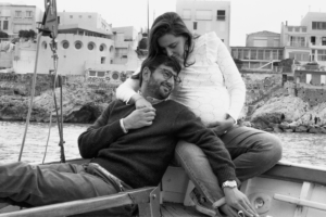 Cette image décris un jeune couple l'un assis, l'autre jambe allongé sur le bord arrière d'un pointu en bois. Ils attendent un heureux évènement comme l'atteste le ventre bien rond sur lequel le jeune appuie son visage.